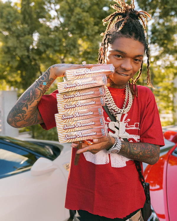 Swae Lee posing with Loose Leaf Sweet Aromatic in front of two sports cars, one beige and one red.