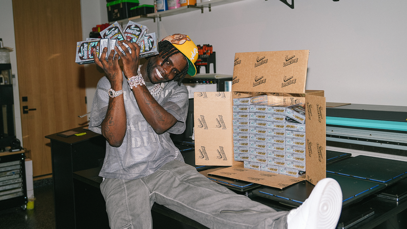 CHIEF KEEF sitting on a table surrounded by ALMIGHTY LEAF Loose Leaf in a warehouse setting