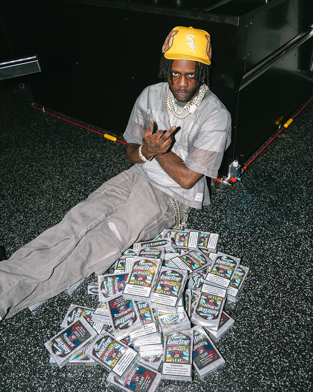 CHIEF KEEF sitting on a table surrounded by ALMIGHTY LEAF Loose Leaf in a warehouse setting