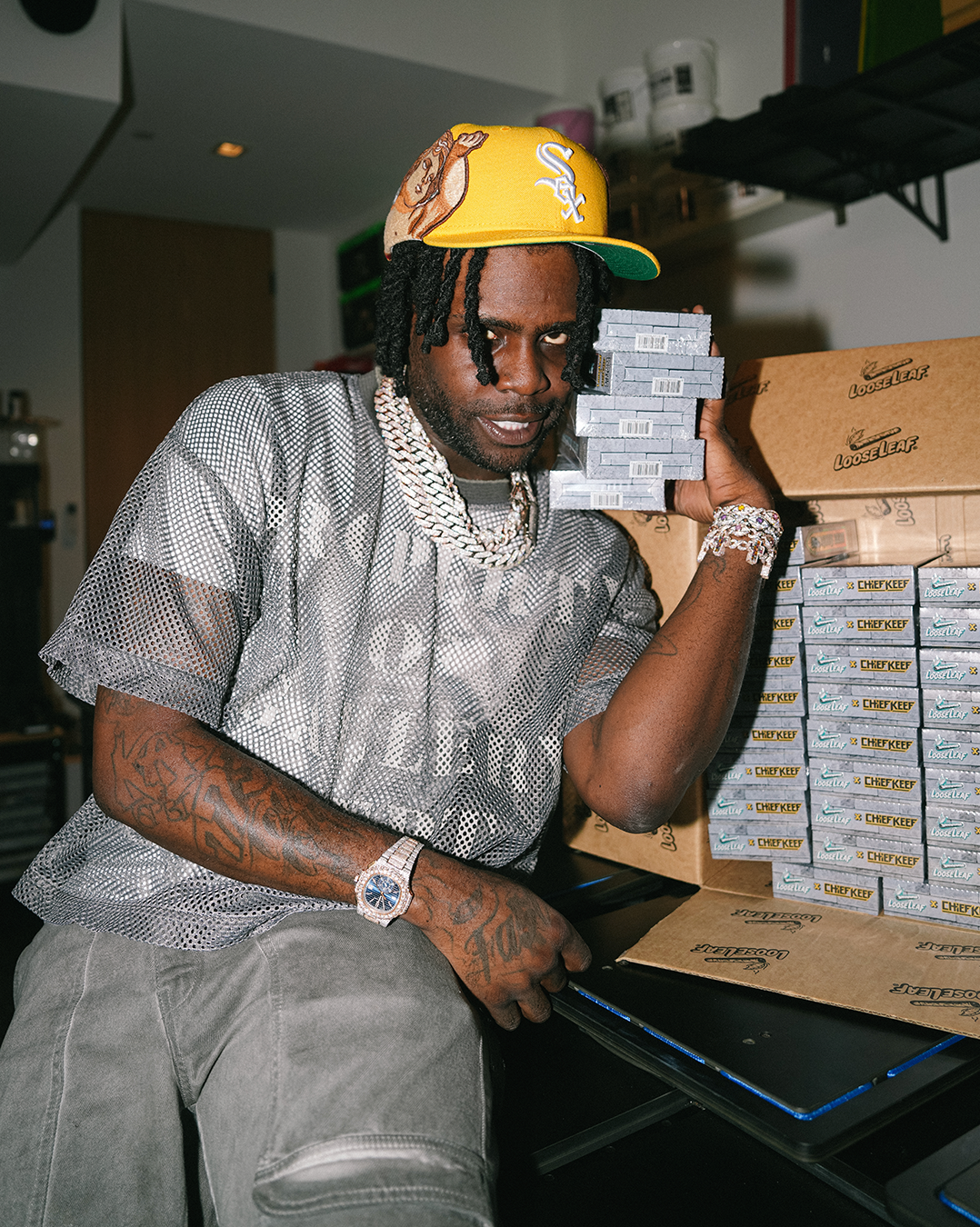CHIEF KEEF sitting on a table surrounded by ALMIGHTY LEAF Loose Leaf in a warehouse setting