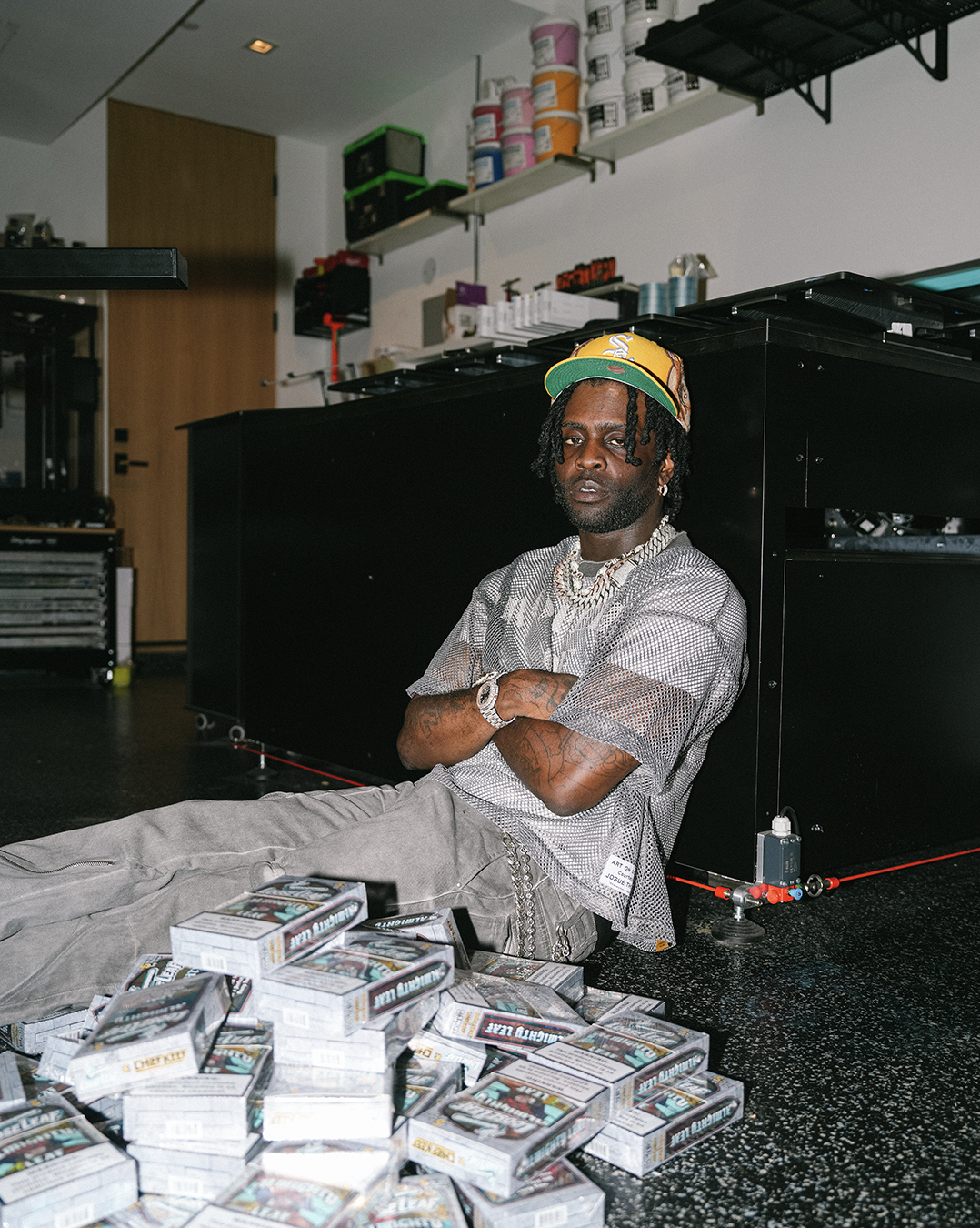 CHIEF KEEF sitting on a table surrounded by ALMIGHTY LEAF Loose Leaf in a warehouse setting