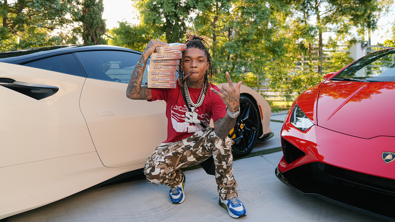 Swae Lee posing with Loose Leaf Sweet Aromatic in front of two sports cars, one beige and one red.